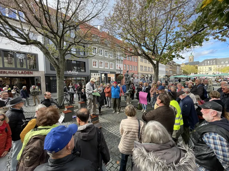 Demonstrierende auf dem Marktplatz unter Bäumen, im Hintergrund Geschäfte