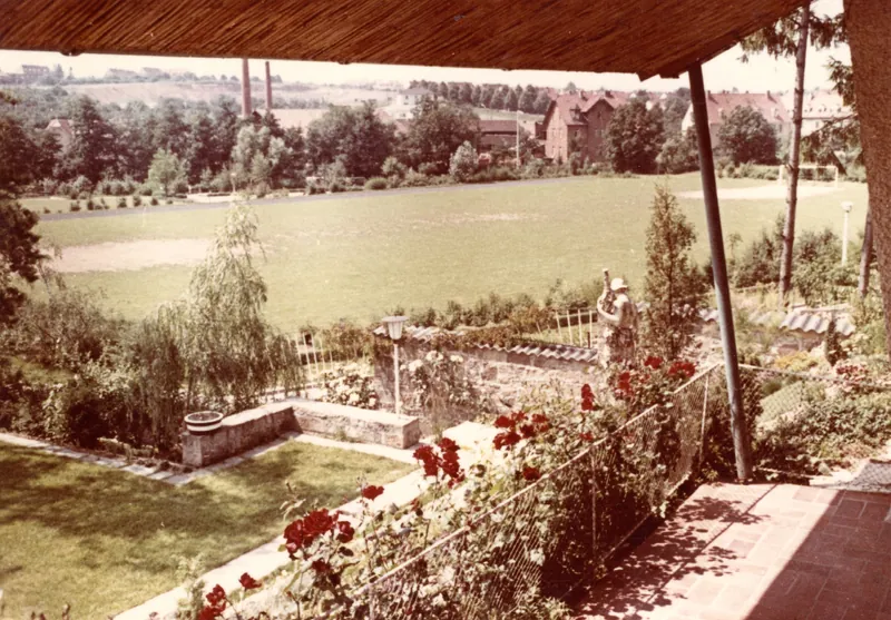 Historisches Farbfoto: Blick von einer Gartenterrasse mit Rosen und Steinmauer über einen großen Naturrasen-Sportplatz. Dahinter Bäume, Backsteinhäuser und zwei Fabrikschornsteine, am rechten Rand ein Fußballtor.
